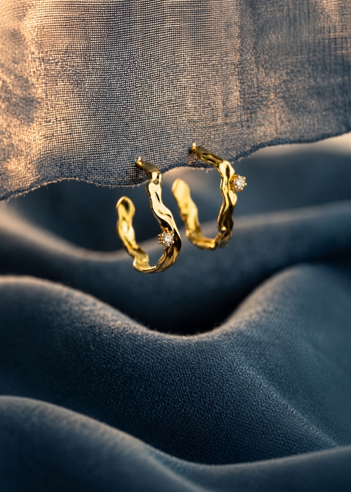 Gold Wave Huggie earrings resting on blue fabric beside wood